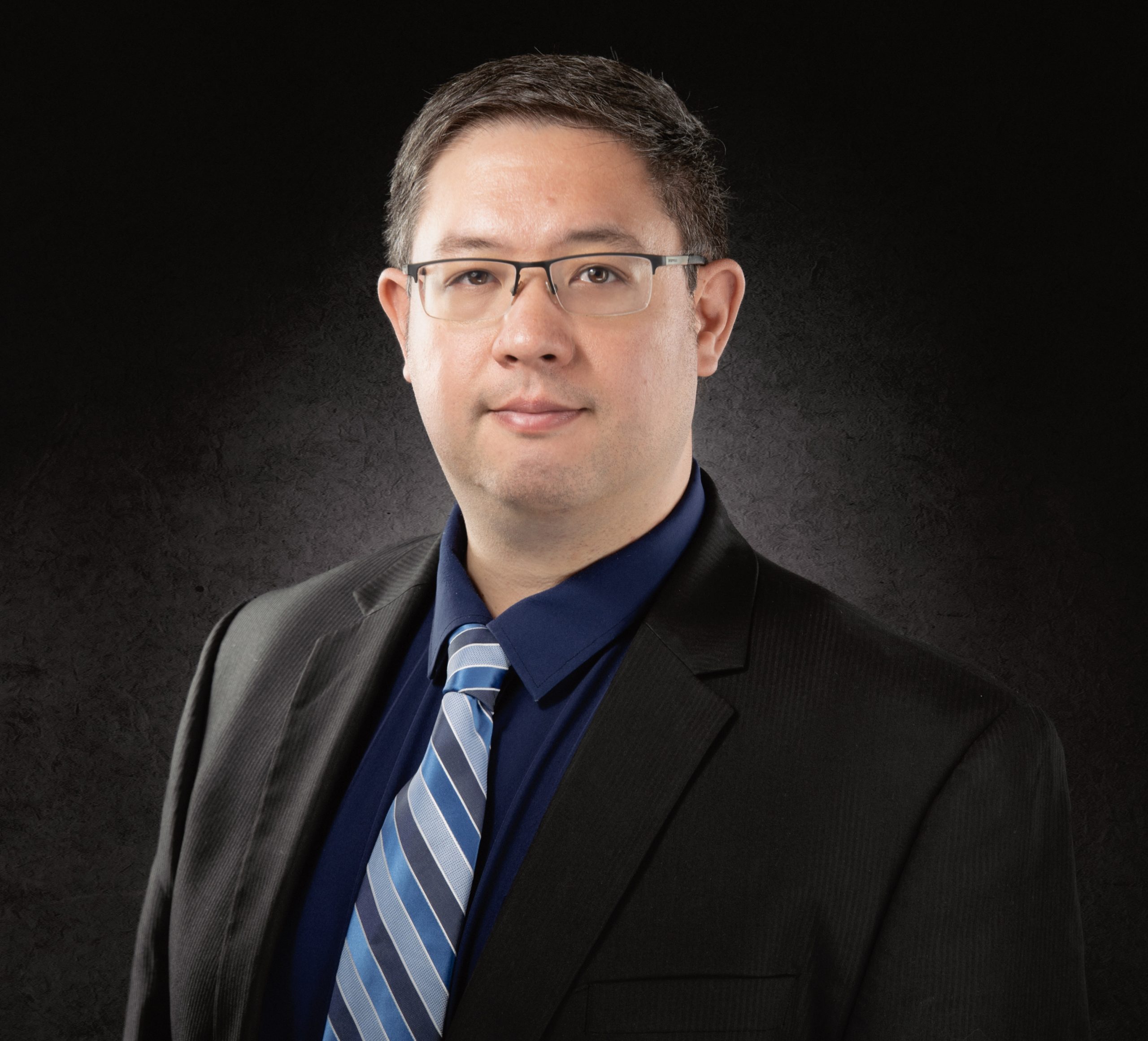 Close-up portrait of a man in a dark suit, blue shirt, and striped blue tie against a black textured background, wearing glasses.