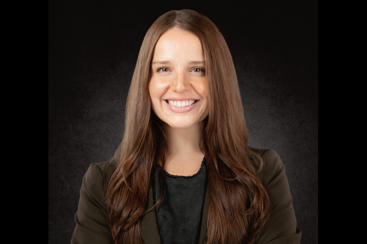 MARIA B HEADSHOTS IMM LAW02077 copy 1 | Richards and Jurusik | Immigration Lawyers | Buffalo, NY Young woman with long brown hair smiling, wearing a dark blazer against a dark studio background.